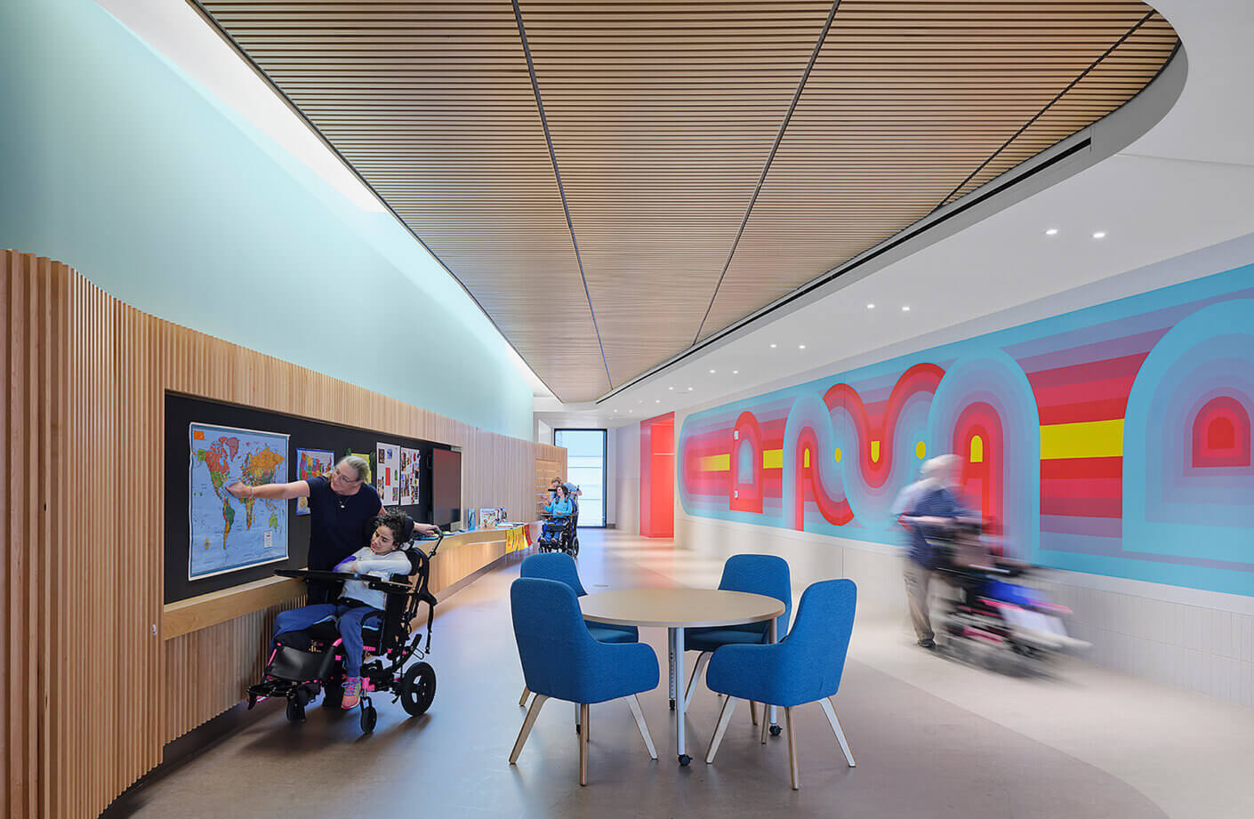 Interior of a bright facility featuring a colorful mural, a map display, and a table with blue chairs. People interact and move throughout the space.