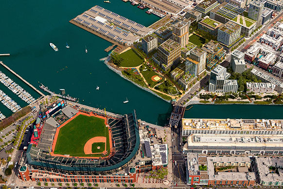 Aerial view of a waterfront area featuring a baseball stadium, residential buildings, a marina, and landscaped parks along the water's edge.