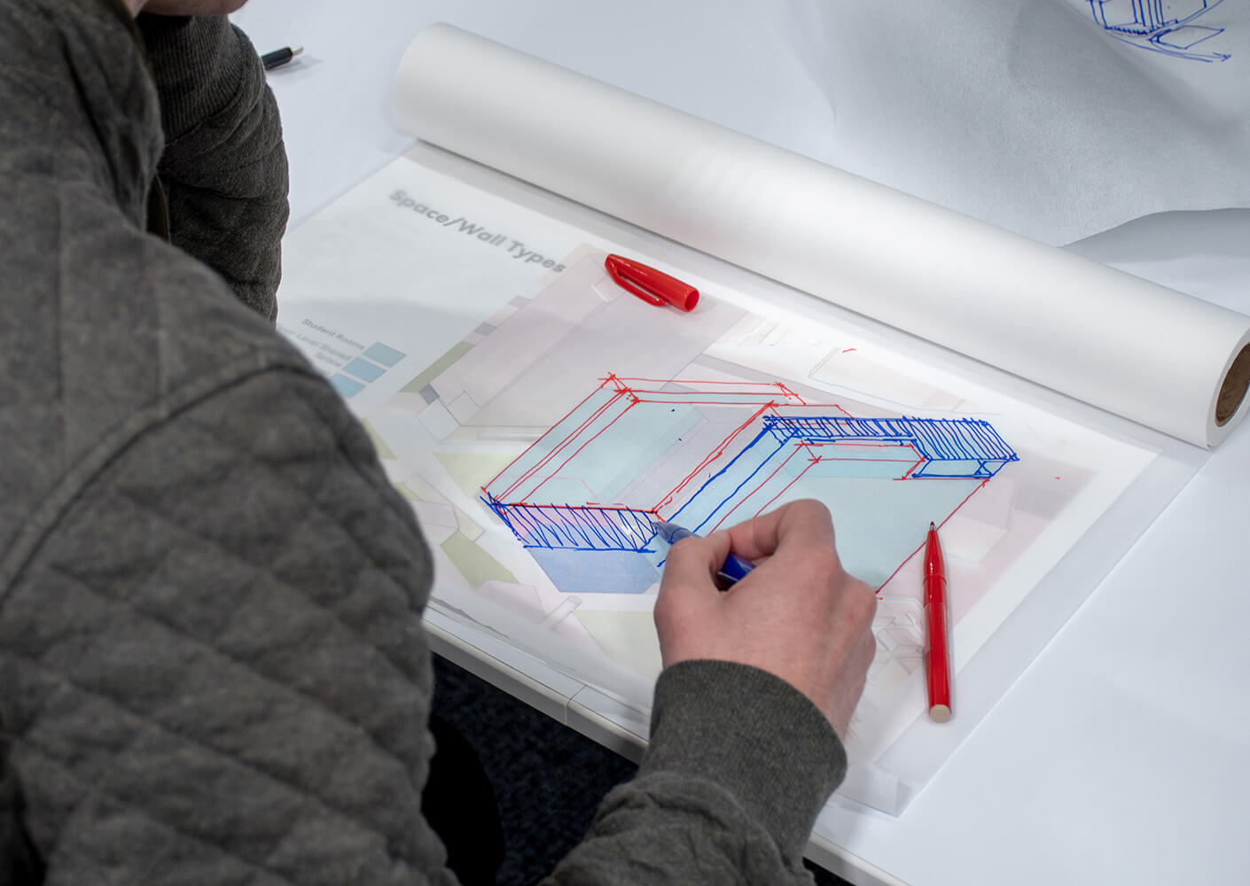 Person sketching architectural designs in blue and red on transparent paper.